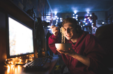 Wall Mural - Happy young man uses a computer at home and eats noodles. Gamer plays video games on a computer at night, holds a plate in his hands, looks at the noodle and smiles.