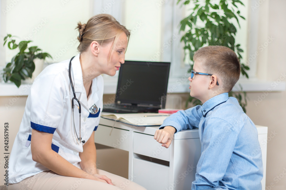 Lekarka w białym kitlu i stetoskopem na szyi siedzi w gabinecie lekarskim. Rozmawia z chłopcem w niebieskiej koszuli i okularach.  - obrazy, fototapety, plakaty 