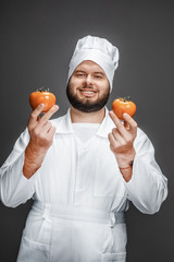 Poster - Bearded guy in chef uniform smiling and demonstrating two ripe tomatoes while standing on gray background