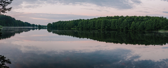 Lake with spring trees panorama photo. Tranquil landscape at a lake, with the vibrant blue sky, white clouds and the trees reflected symmetrically in the clean blue water.