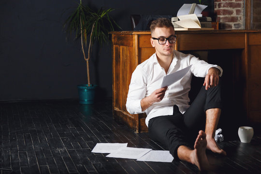 Handsome writer man wearing glasses and white shirt sitting on the floor near the table with typewriter, drinking coffee and reading new chapter of his book