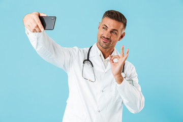 Canvas Print - Photo of kind medical doctor wearing white coat and stethoscope taking selfie on smartphone, standing isolated over blue background