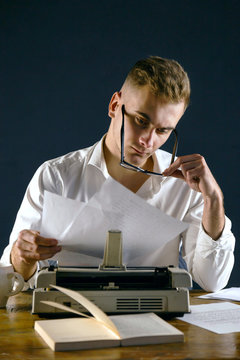 Handsome writer man wearing glasses and white shirt sitting at the table with typewriter and looking under the glasses on the new pages of a his book.