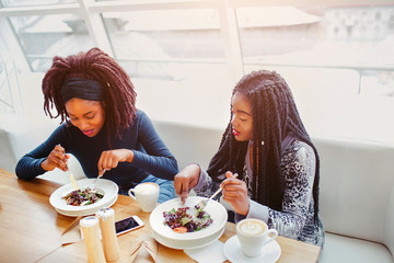 Two african women sit at table and eat food. They concentrated. Models are in cafe. Thy devour meal.