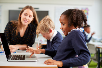 A female school teacher sitting at a desk helping a boy and girl using a laptop computer in primary school class, side view