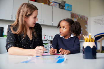 young female primary school teacher working one on one with a schoolgirl at a table in a classroom, 