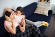© estradaanton - Sick young woman sit on couch together with her daughter. Child embrace mother head. She look at her. Kid touch woman's hand. She try to comfort her.