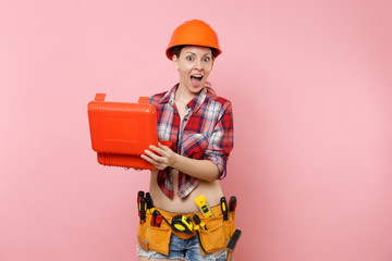 Wall Mural - Strong young handyman woman in orange helmet, plaid shirt, denim shorts, kit tools belt full of instruments, toolbox isolated on pink background. Female in male work. Renovation and occupation concept