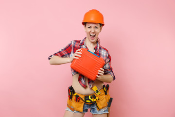 Wall Mural - Strong young handyman woman in orange helmet, plaid shirt, denim shorts, kit tools belt full of instruments, toolbox isolated on pink background. Female in male work. Renovation occupation concept