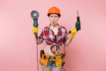 Wall Mural - Energy fun handyman woman in gloves, protective orange helmet, kit tools belt full of instruments holding power saw electric drill isolated on pink background. Female in male work. Renovation concept