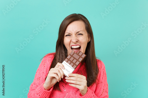 Fotografie, Tablou Cheerful young girl in knitted pink sweater holding in hand, biting and eating chocolate bar isolated on blue turquoise wall background, studio portrait