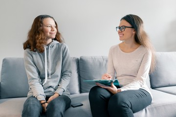 Young woman professional psychologist talking with teenager girl 14, 15 years old sitting in office on sofa. Mental health of child in adolescence