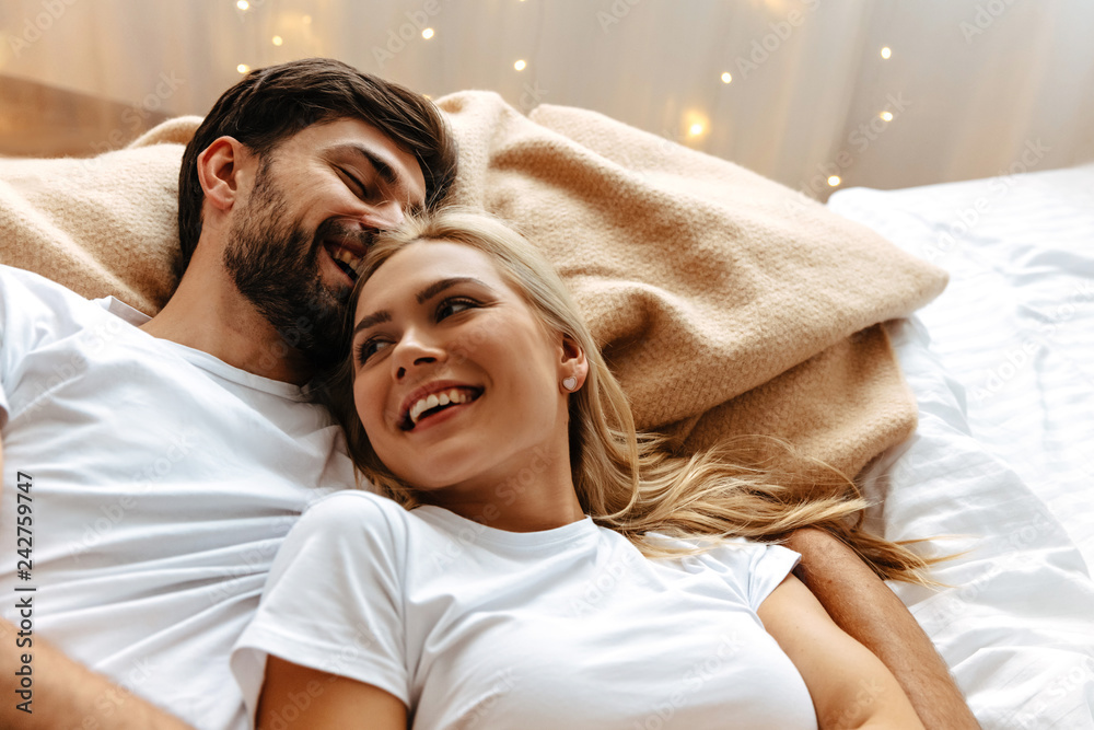 Love. Fun. Emotions. Young couple are laughing while lying together on the bed - obrazy, fototapety, plakaty 