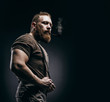 Lumberjack brutal red beard muscled man in brown shirt with smoking tube standing on dark background. Handsome man with red beard and moustache smoking pipe