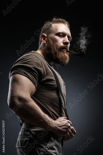 Fotografía Lumberjack brutal red beard muscled man in brown shirt with smoking tube standing on dark background