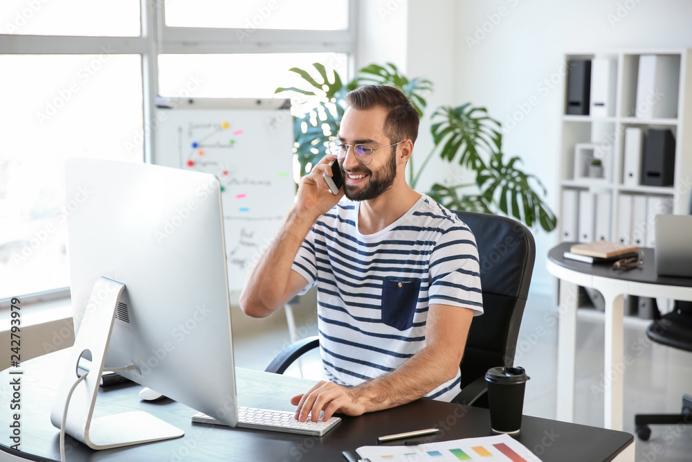 Young man talking on mobile phone while working on computer in office