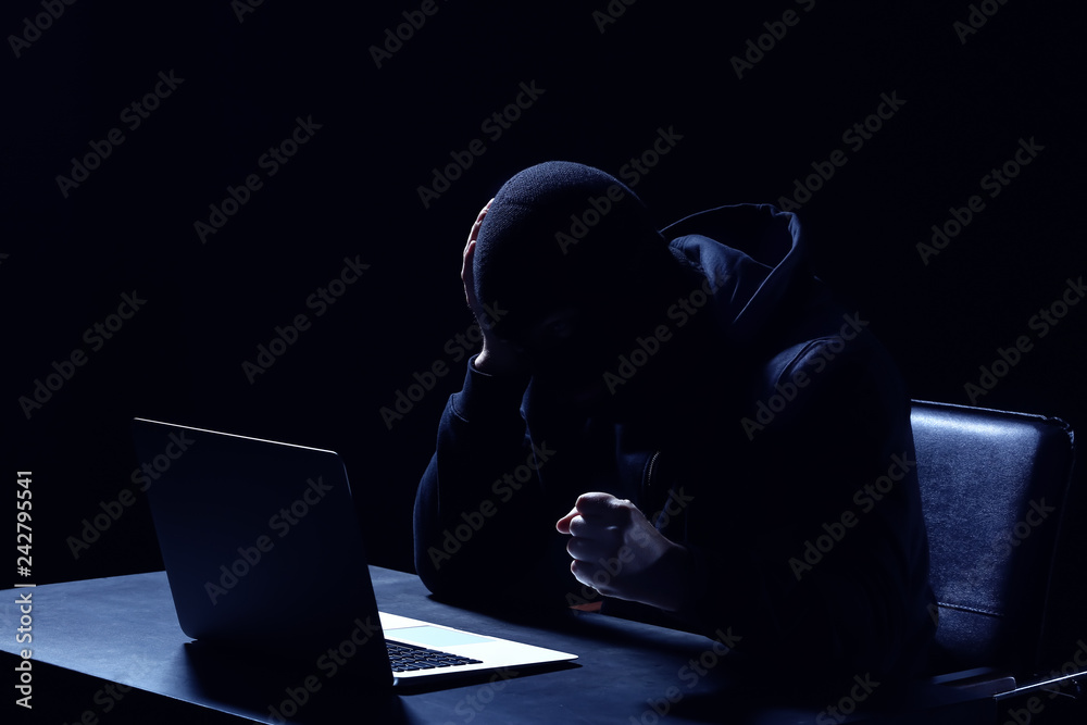 Emotional hacker with laptop sitting at table on dark background