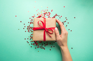 Wall Mural - Gift box wrapped in brown colored craft paper and tied with red bow on blue background with red confetti. Lady's hand holding a giftbox.