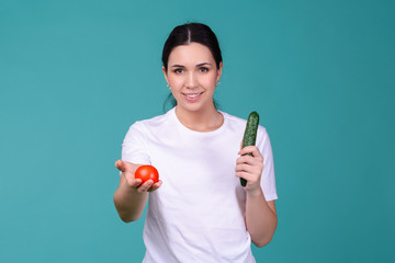 Wall Mural - Young attractive woman in t-shirt holding ripe juicy tomato and big green cucumber in her hands and giving it to you. Healthy vegetarian food concept. Spring Avitaminosis prevention.