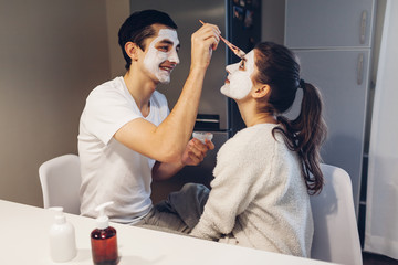 Wall Mural - Man applying clay mask on his girlfriend's face. Young loving couple taking care of skin at home