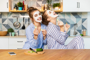 Portrait of two best friends laughing and having fun on kitchen with nice interior. Cheerful models wearing face masks. Friendship and cleansing concept. Blurred background