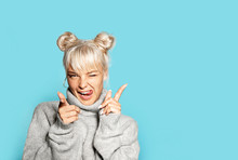 Portrait Of Beautiful Blonde Girl Wearing Cozy Grey Sweater And Making Faces. Pretty Model In Cheerful Mood Posing On Blue Background. Copy Space In Right Side. Winter And Emotion Concept