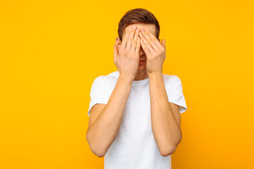 Portrait of a frightened man with glasses, the guy closed his eyes with his hands for fear, on a yellow background