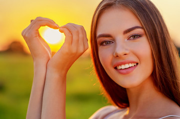 Wall Mural -  Pretty joyful  girl gesturing heart shaped over sunset background.