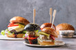 Variety of homemade classic and mini burgers in wheat and black buns with beef and veal cutlets, melted cheese and vegetables on white ceramic board over grey blue table.