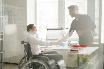 Businessman shaking hands with disabled man in wheelchair behind the window at office