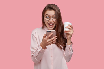 Wall Mural - Impressed beautiful woman feels overjoyed, reads text message from excitement, dressed formally, carries takeaway coffee, isolated over pink studio wall. People, communication, lifestyle concept