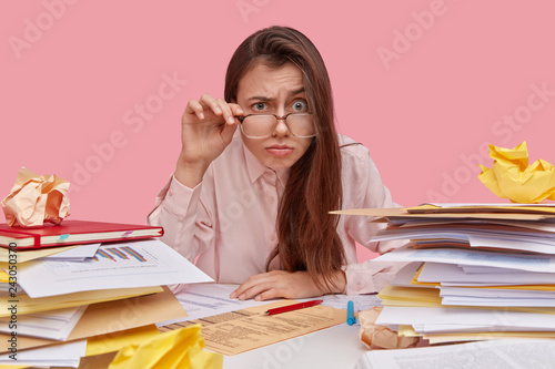 Fotografía Photo of funny female wonk looks through thick lenses of spectacles, has surprised indignant expression, much paper work, heap of documents on desk, isolated over pink background