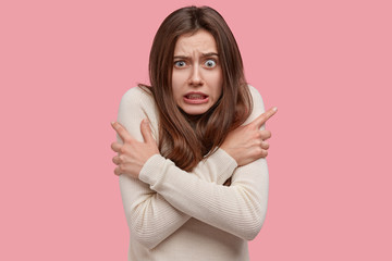 Canvas Print - Photo of dissatisfied beautiful woman crosses arms over chest, trembles from cold, frowns face and clenches teeth, stands over pink background, afraids of something terrible. Negative feelings