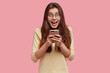 © WHstudio Leushin N - Waist up shot of pretty satisfied woman holds mobile phone, happy to exchange text messages in social networks, wears yellow jumper, models over pink background. Online communication concept