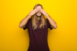 Blond man with long hair over yellow wall covering eyes by hands. Surprised to see what is ahead