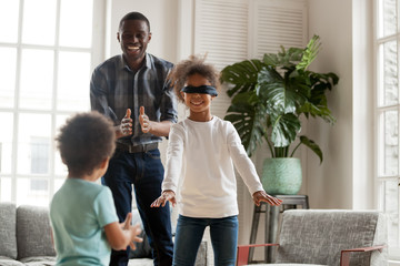 Blindfolded little girl playing hide and seek at home with african dad and kid brother, happy black dad and 2 mixed race children laughing enjoy active game together, family having fun in living room