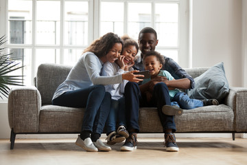 Wall Mural - Happy african family with 2 children son daughter having fun with gadget on couch using smartphone at home, black parents and kids laugh watch funny video, make video call selfie on phone online app