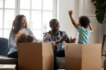 Excited mixed race kids jumping out of box playing with mom dad in living room, african joyful children having fun laughing packing unpacking with black parents, family moving in new home relocation