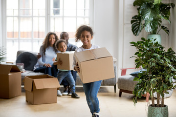 Wall Mural - Happy mixed race kids running carrying boxes on moving day, black family tenants relocation concept, african american parents and children laughing playing in new home flat having fun in living room