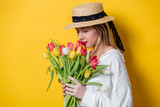 Fototapeta Tulipany - Beautiful woman in white shirt and hat with fresh springtime tulips on yellow background