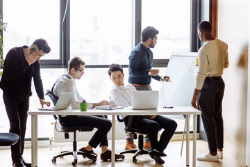 Wall Mural - Multiethnic group of young business men in trendy office clothing negotiating standing near large panoramic window with big cityscape, partners having business discussion in contemporary office