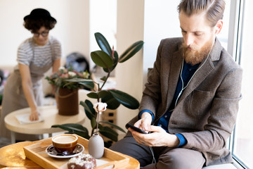 Wall Mural - Serious concentrated hipster bearded man in earphones sitting on window-sill and surfing net on modern gadget while spending morning in cafe