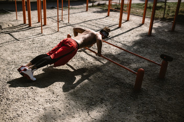 Wall Mural - Young athletic man in headband  with a naked torso with tattoos dressed in black leggings and red shorts doing push-ups on the uneven bars outside on a sunny day