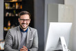 © eshana_blue - Portrait of handsome businessman smiling at camera while sitting in front of computer at the office desk