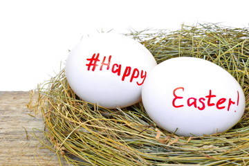 Two chicken eggs in straw nest with text happy Easter and sign hashtag for social network on wooden rough board on white background. Close up