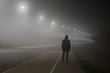 © fotoduets - Young man alone slowly walking under white street lights in night. Dark time. Peaceful atmosphere in mist. Foggy air. Back view.