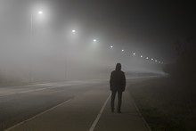 Young Man Alone Slowly Walking Under White Street Lights In Night. Dark Time. Peaceful Atmosphere In Mist. Foggy Air. Back View. 