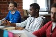 © pressmaster - Young African-america specialist sitting by table between colleagues and making organization notes