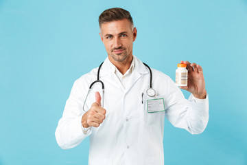 Canvas Print - Mature man doctor posing isolated over blue wall background holding pills vitamins showing thumbs up.