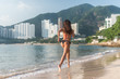 Back view of fit slim girl running barefoot on seashore wearing bikini. Young woman doing cardio exercise beach lit in sunshine and city mountains in background.